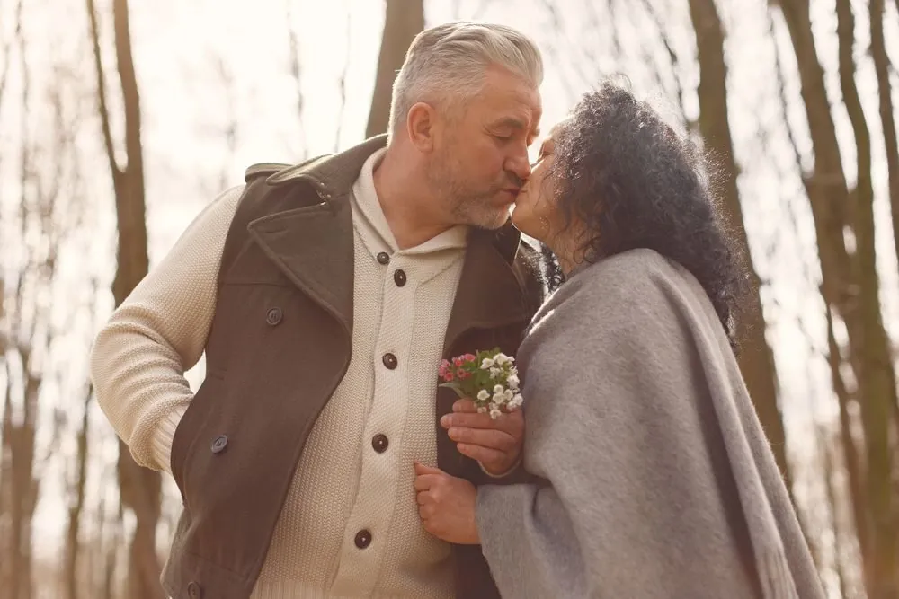 Senior couple sharing a tender kiss in a sunlit forest, expressing love and affection.