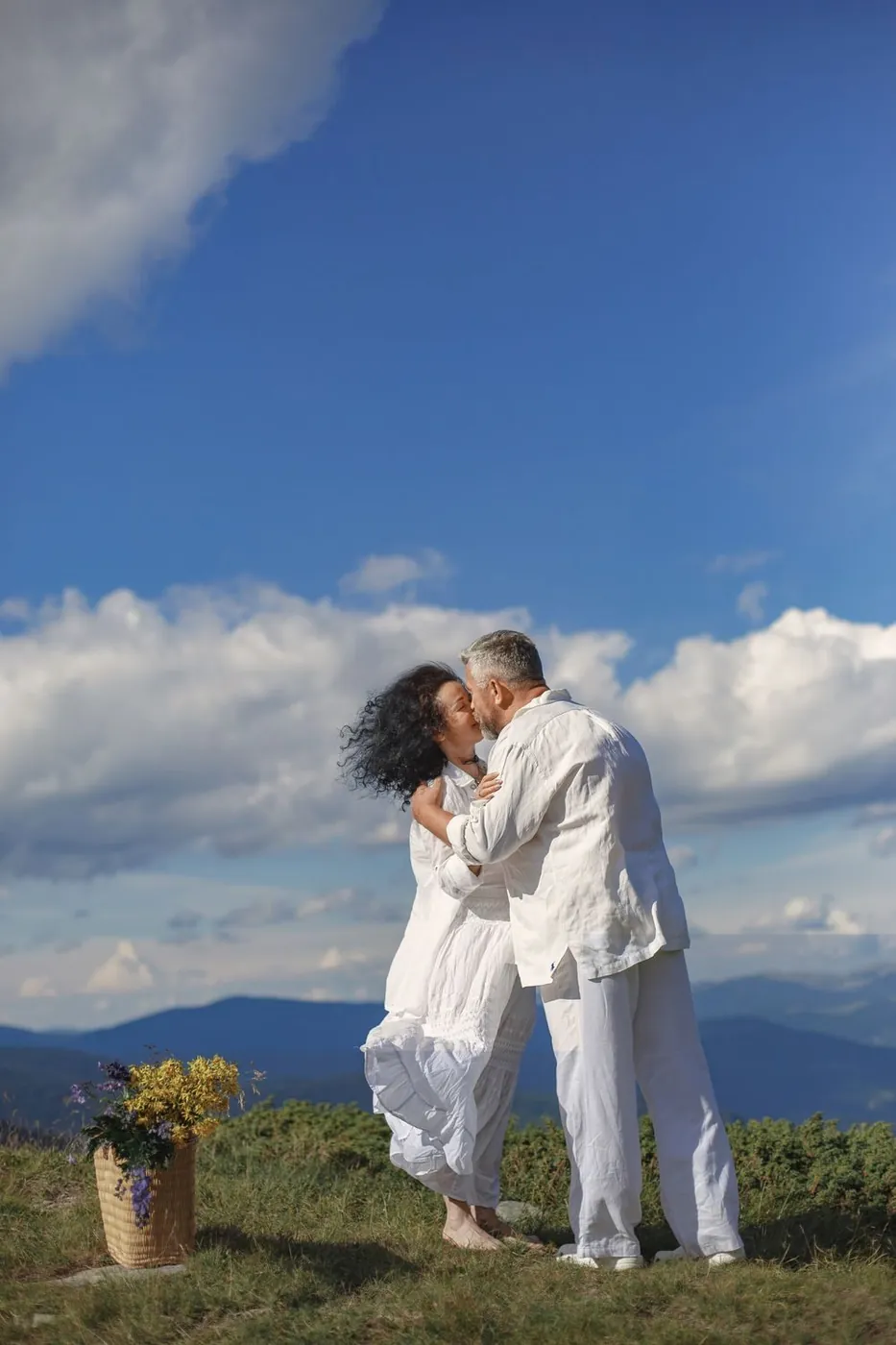 Couple in white clothing shares a romantic kiss on a hilltop with a blue sky backdrop.
