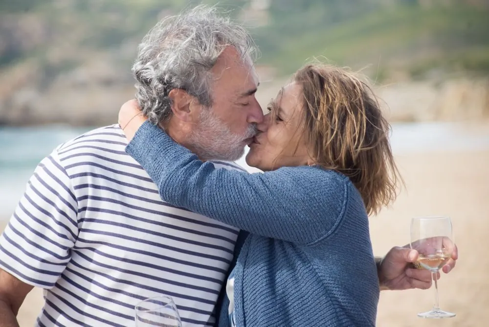 Seniors enjoying a romantic moment on a beach in Portugal, holding wine glasses.