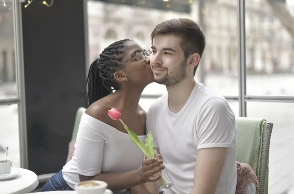 A multiracial couple shares a tender kiss while enjoying a coffee indoors, symbolizing love and intimacy.