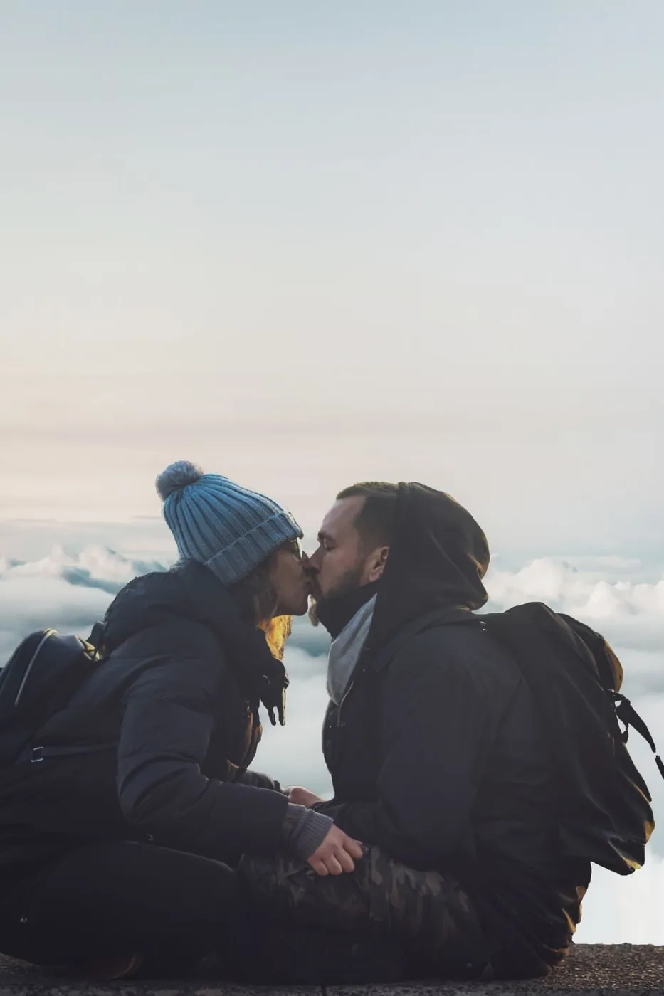Romantic couple kissing in winter clothing on a mountain top with a misty sky backdrop.