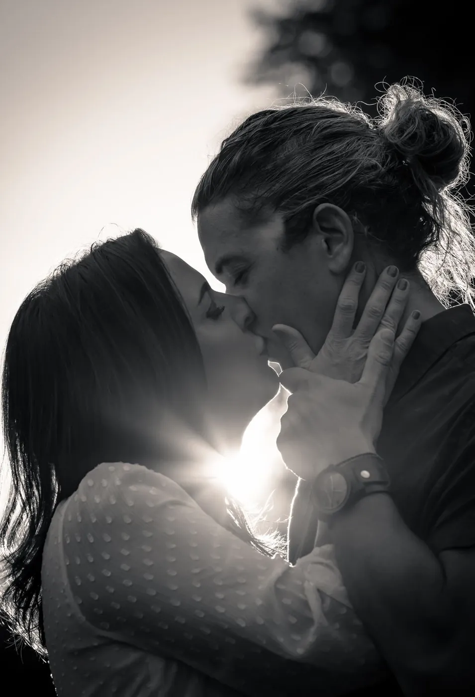 A black-and-white photo capturing a romantic moment between a couple kissing outdoors with sunlight in the background.