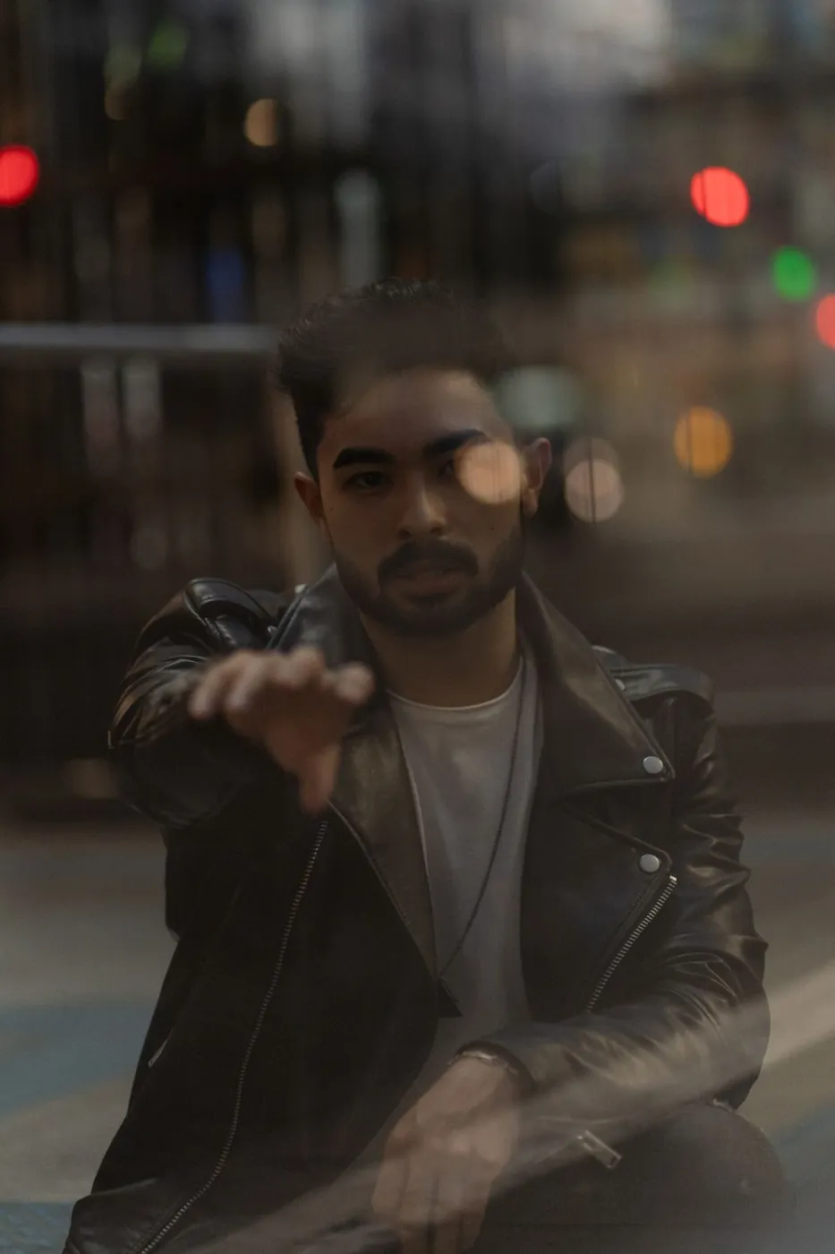 A man in a black leather jacket reaching toward a window, capturing a moody city reflection.