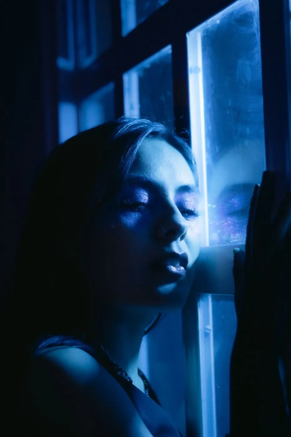 A woman basks in soothing blue light, eyes closed by a window.