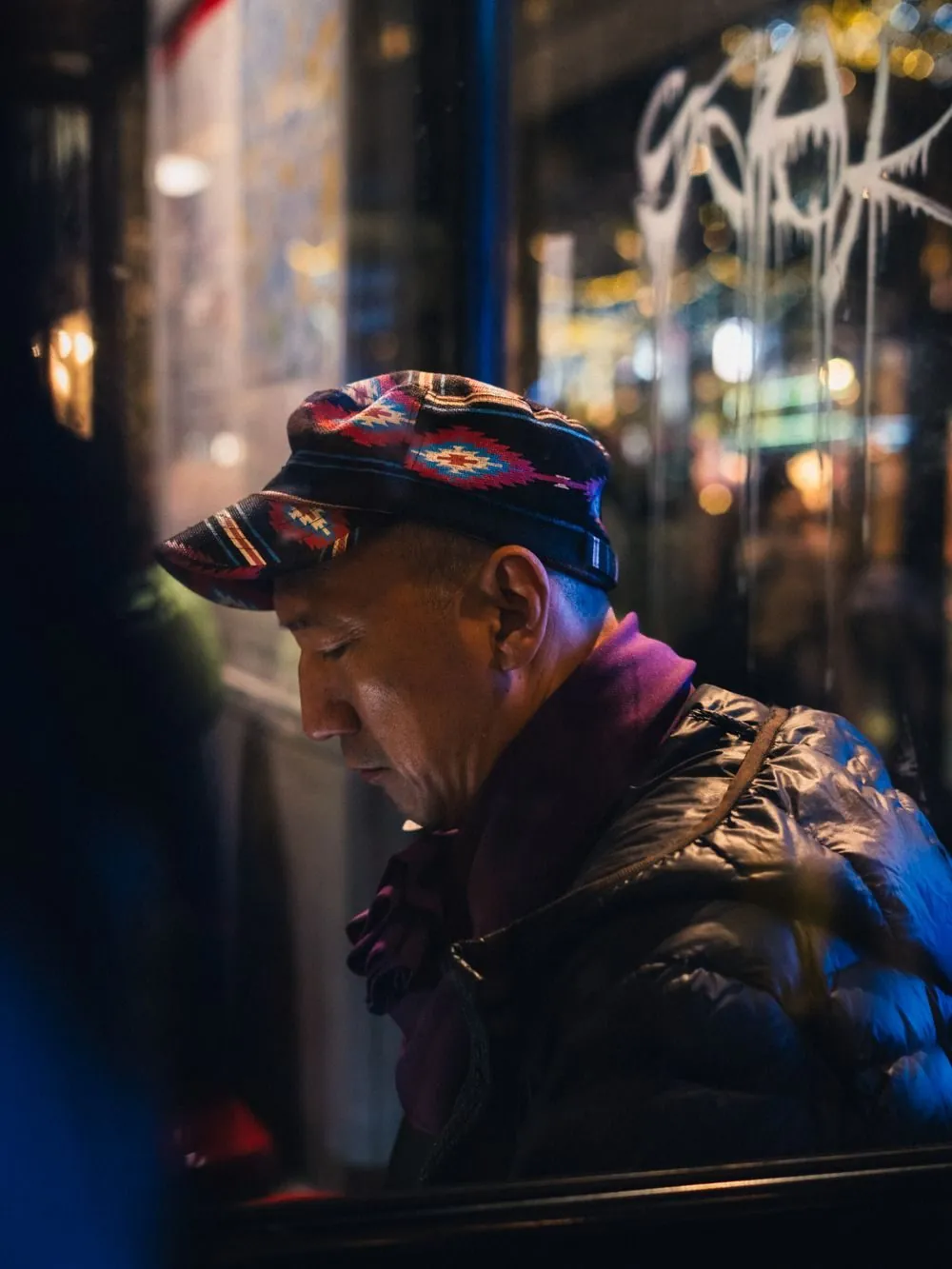 A contemplative man wearing a colorful hat and scarf, captured in a warmly lit urban setting at night.