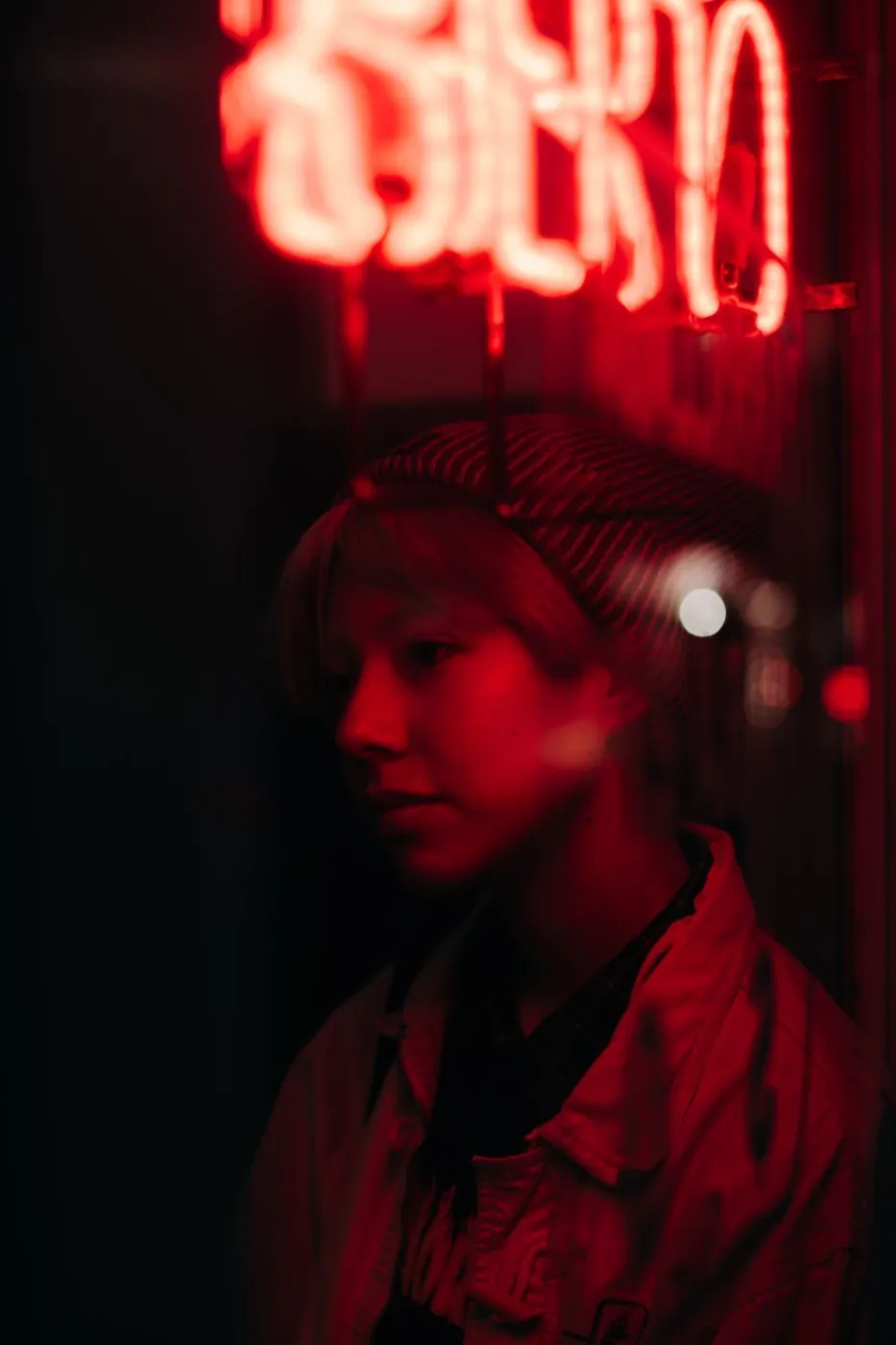 A young woman in a beanie reflects under red neon lights, creating a moody ambiance.