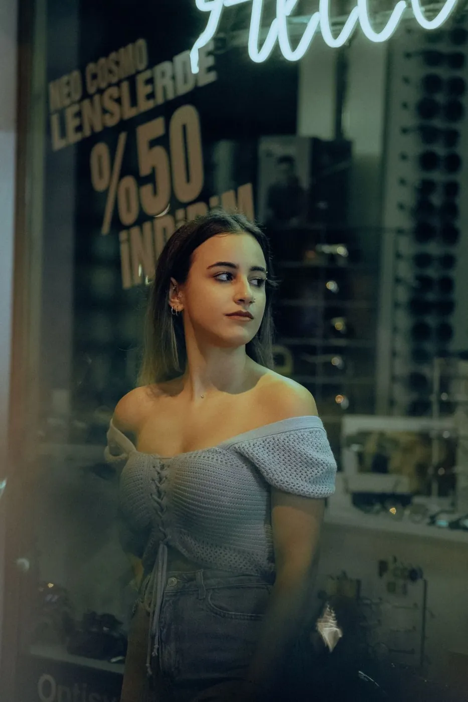 Stylish woman in a crochet top stands by a shop window with neon sign reflections in the evening.