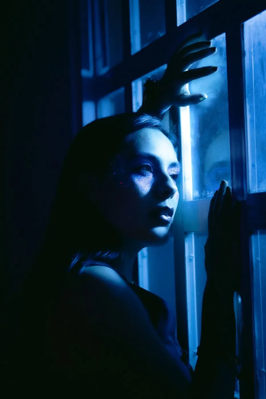 A woman gazes thoughtfully out a window, illuminated by moody blue light.