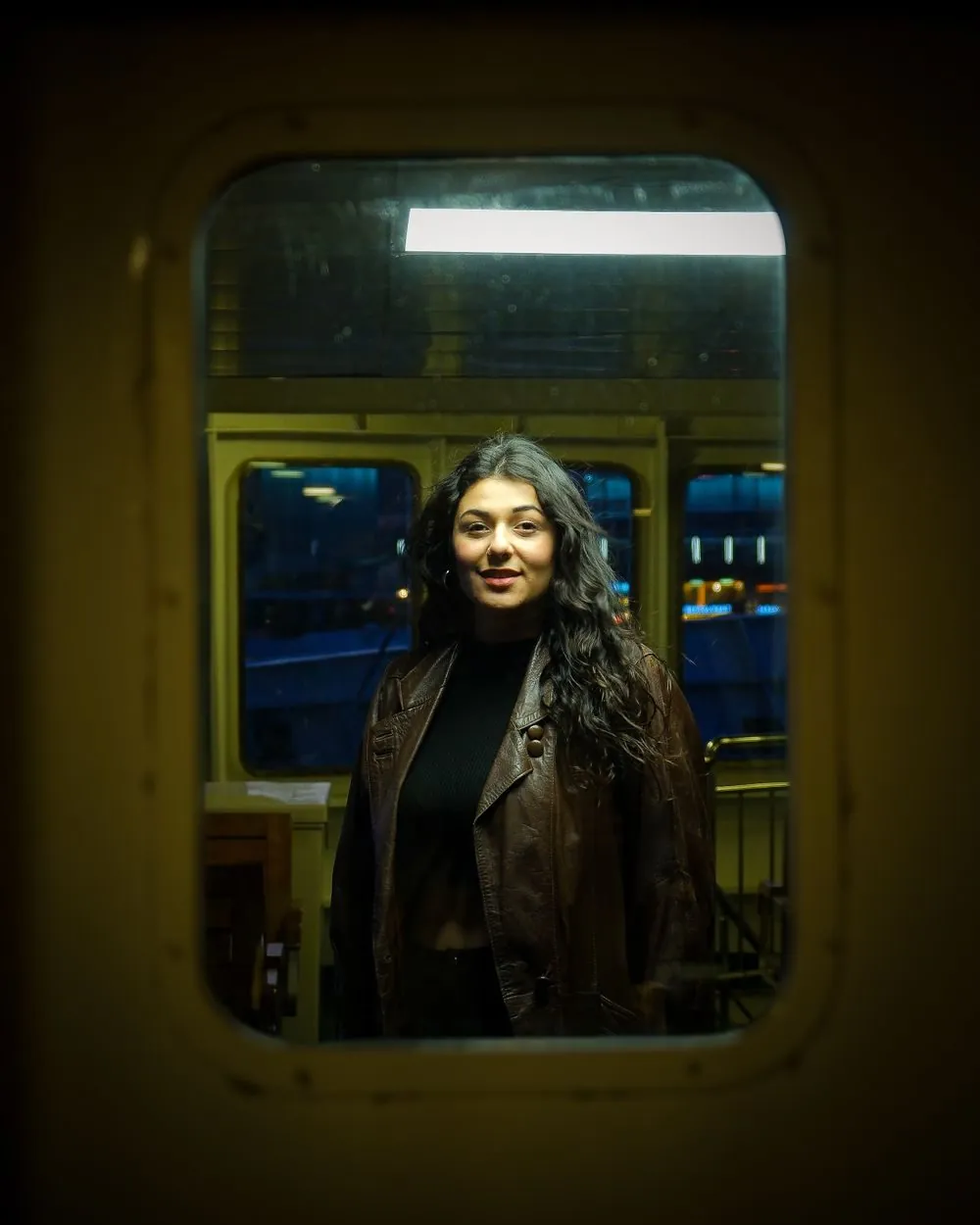 A woman with curly hair in a leather jacket smiling at night on an İstanbul ferry.