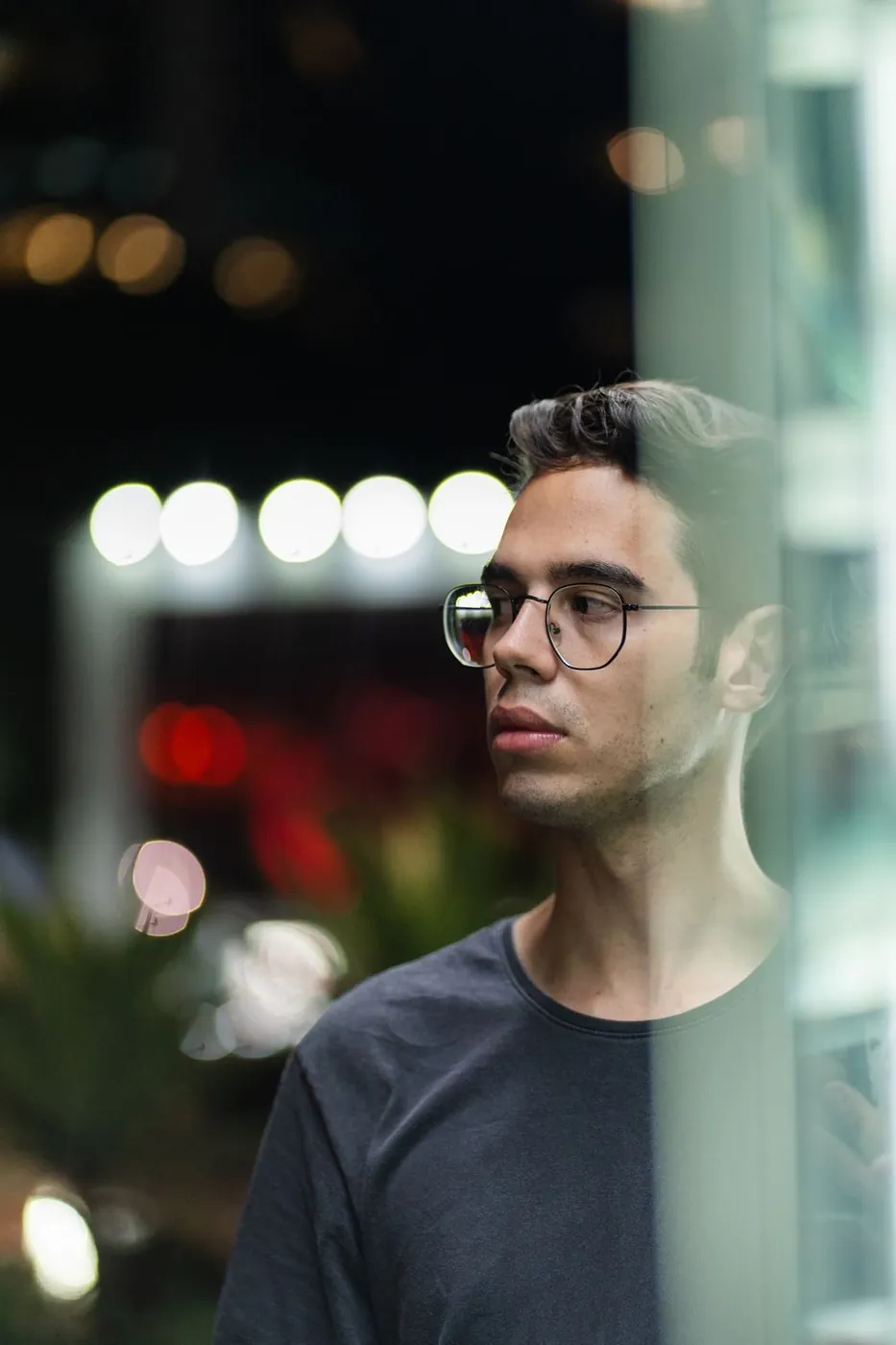 Young man wearing glasses gazes thoughtfully through glass, urban night lights bokeh.