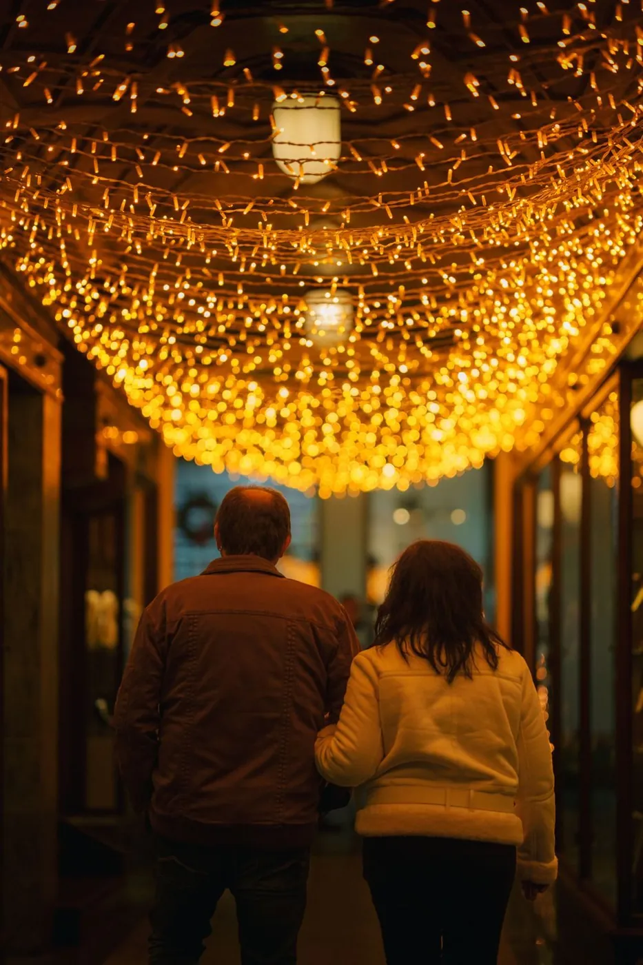 A couple strolls through a festive corridor adorned with warm golden lights.