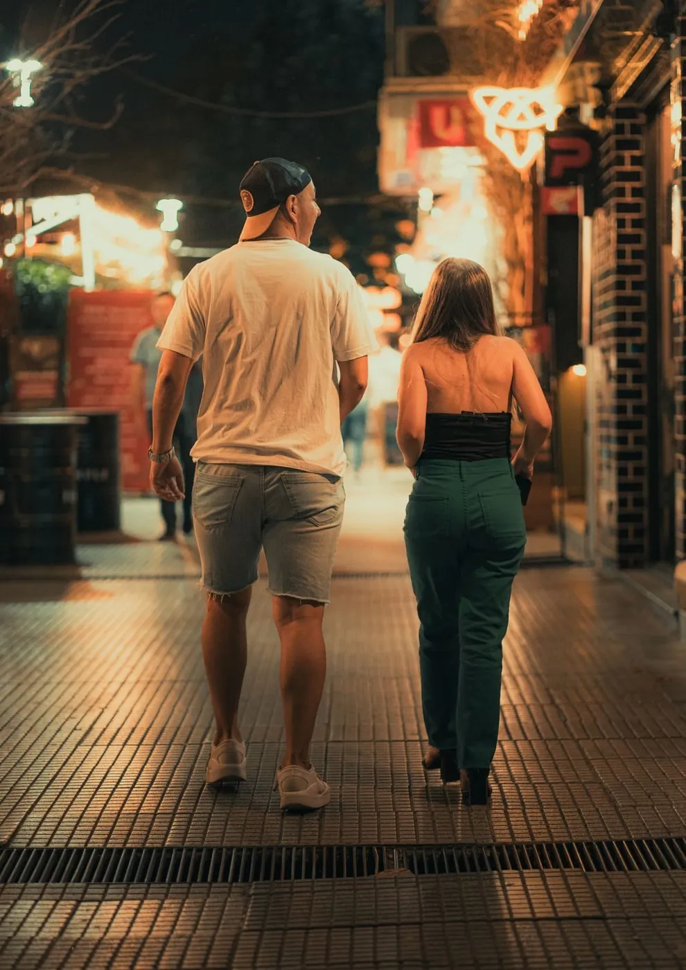 A couple walks down a vibrant street at night, surrounded by glowing lights and lively atmosphere.