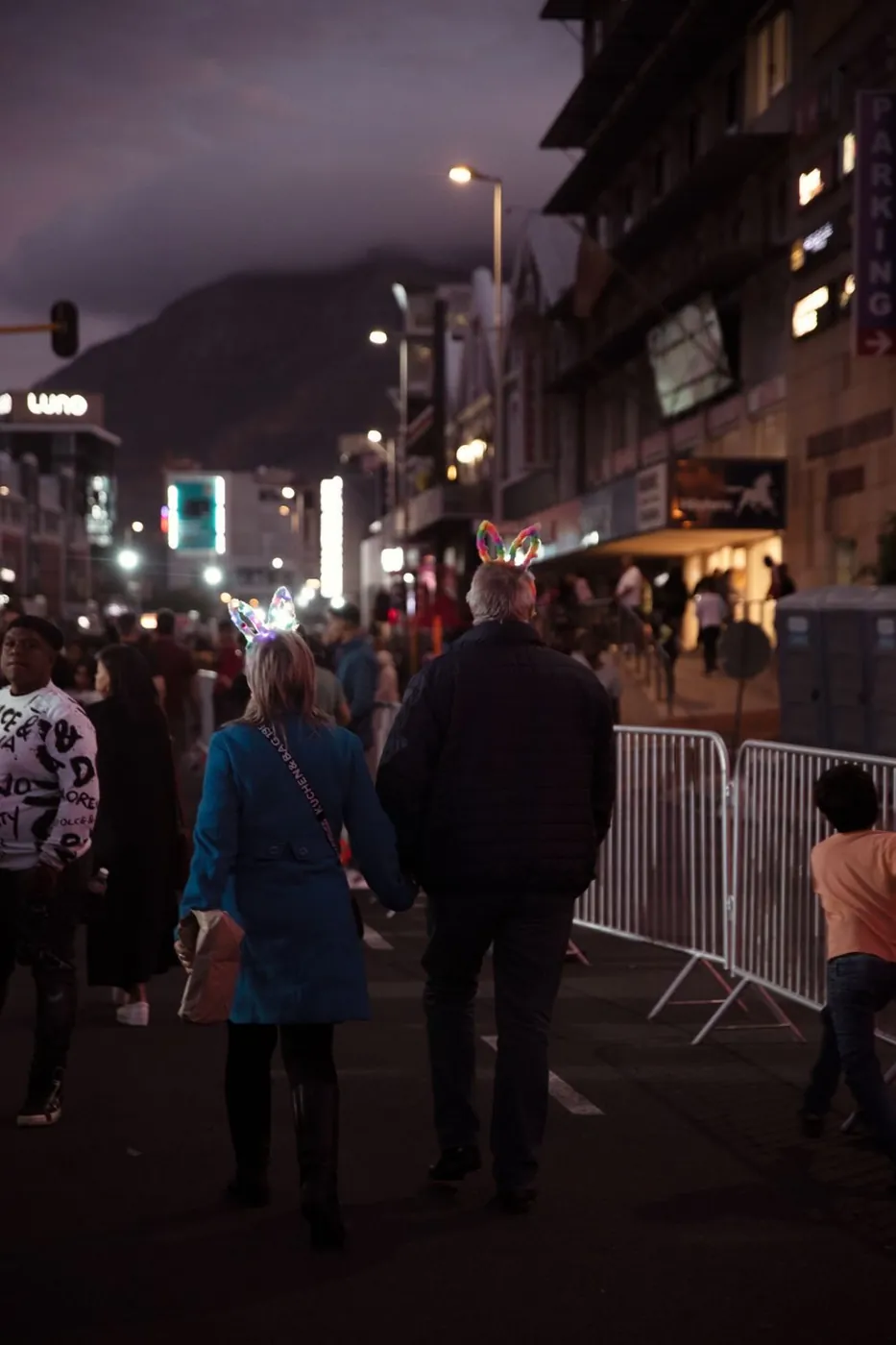 Older couple with bunny ears walking among a city crowd during an evening festival.