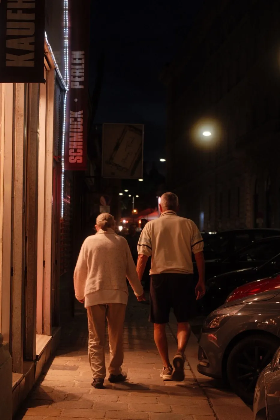 An elderly couple walks hand in hand down a street at night, displaying love and companionship.