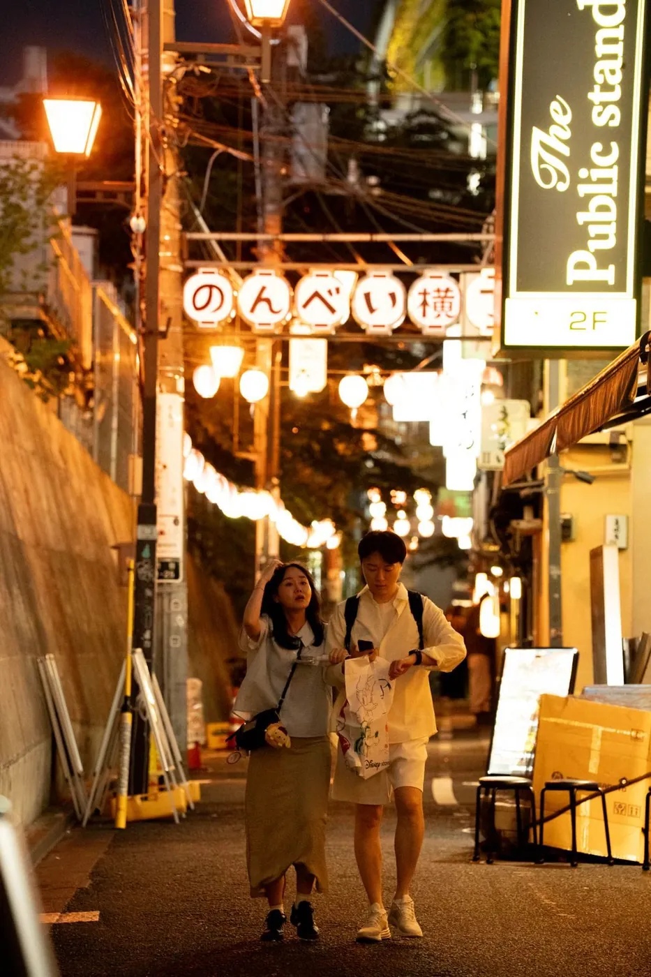 A couple walking through a vibrant Tokyo alleyway at night, surrounded by glowing lanterns and storefronts.