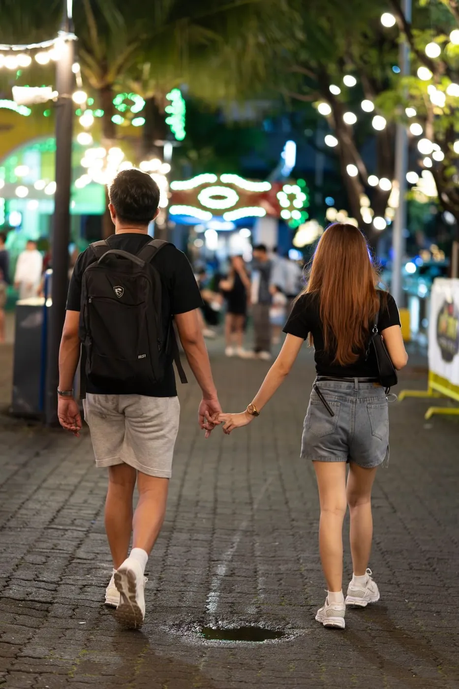 A young couple walking hand in hand on a brightly lit street at night, capturing a romantic city vibe.
