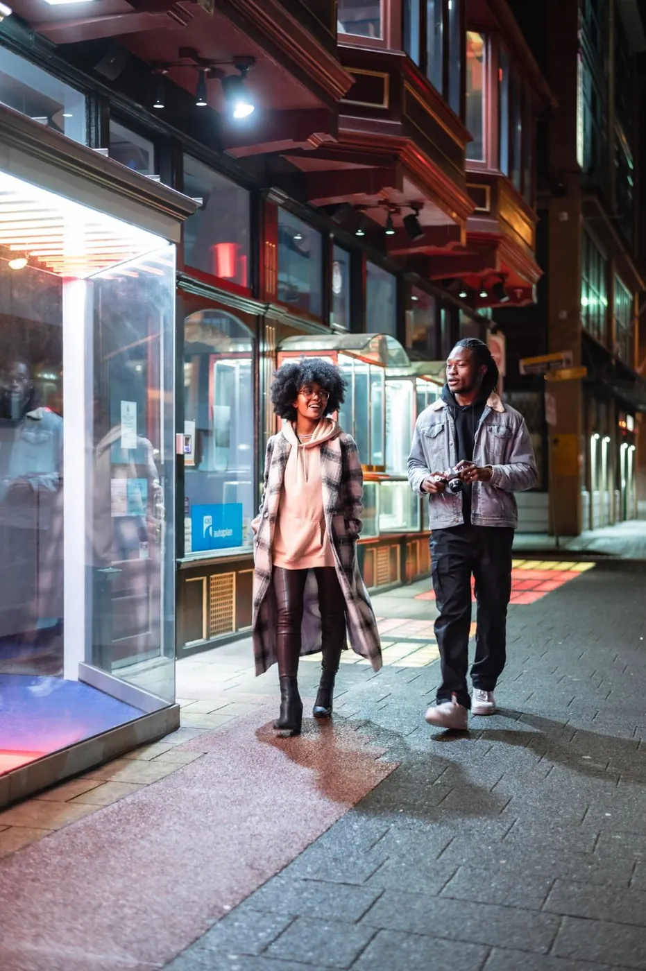 Full body of stylish African American couple with photo camera strolling on paved sidewalk near glass wall of modern building in city
